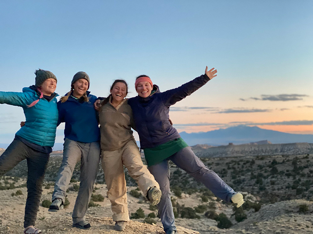 A group of three stand in the desert hugging and facing the camera. They have smiles on their faces and the sun is setting in the background.